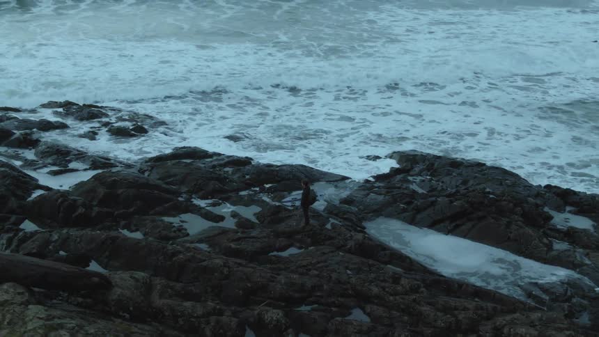 A person dressed in black holding a surfboard is standing on rocks watching the rough water and waves roll in