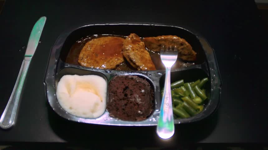 A tv dinner with meat potatoes green beans and a brownie on a black table with a knife and fork