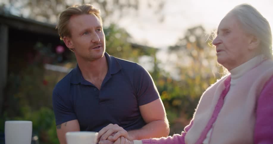 A middleaged man in a tight shirt holds an elderly womans hand in both of his hands as they sit in a sunny backyard and chat