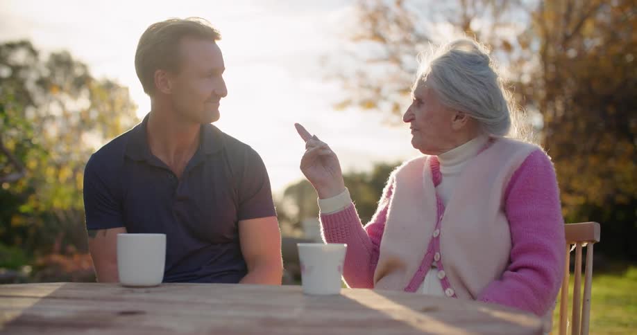 Man wearing a dark blue polo shirt and elderly woman with pink sweater and white vest sit at table and talk to each other in the garden as the sun shines brightly in the background
