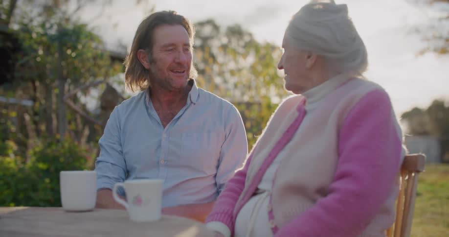 Middle aged man with a light blue shirt and elderly woman wearing a pink and white sweater sit at table and talk to each other in the garden
