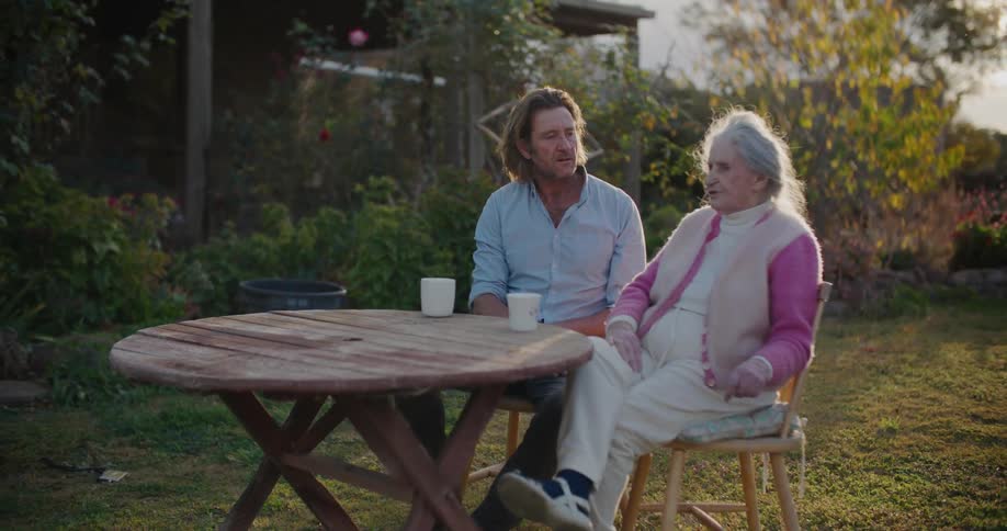 A man chats with his grandmother over coffee in their backyard