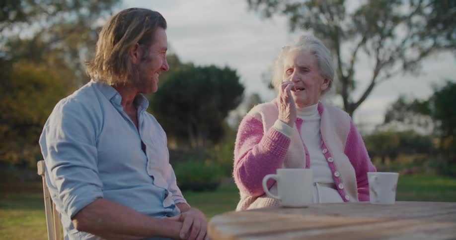A man laughs while listening to his grandmother as they sit outside with coffee mugs