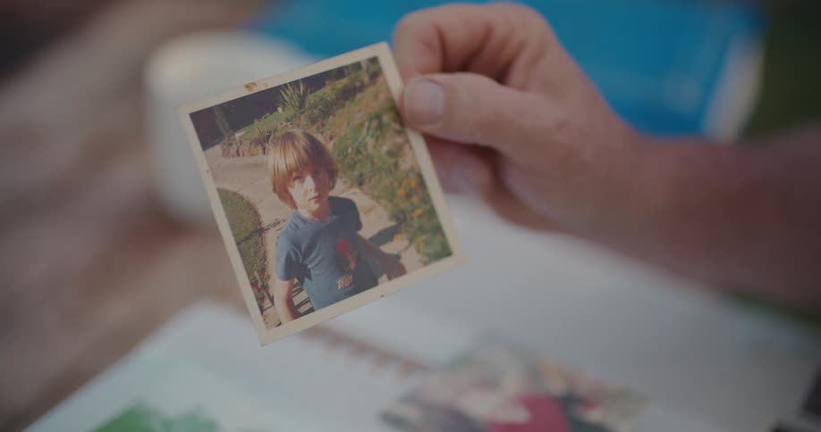 A man holds up a square shaped photograph of a boy standing on a sidewalk