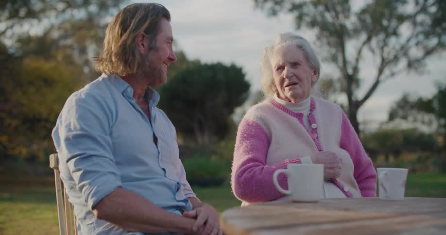 Middle aged man with a light blue shirt and elderly woman sit at table and talk to each other in the garden