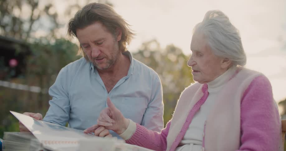 Middle aged man with a light blue shirt and elderly woman with a white vest and a pink sweater look at photo album in the garden