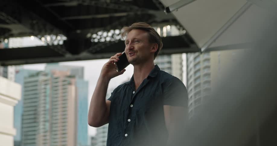 A man happily talks on a smartphone while standing outdoors in a metropolis during the day