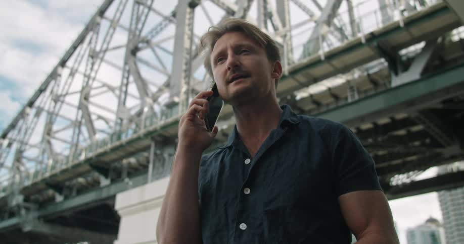 A man talking on a smartphone with a bridge the sky and clouds in the background