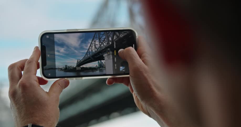 A man with a smart phone taking a picture of a bridge with the sky and clouds in the background