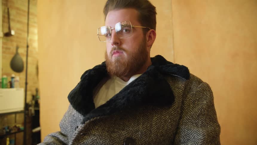 A man is sitting on a chair and wearing a coat and glasses