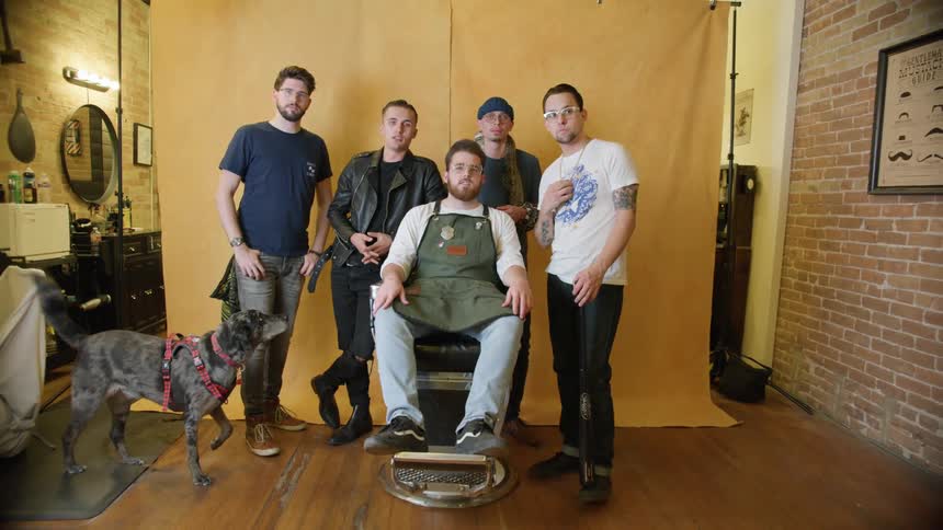 Five men in a barbershop with a dog with a wall and mirror in the background