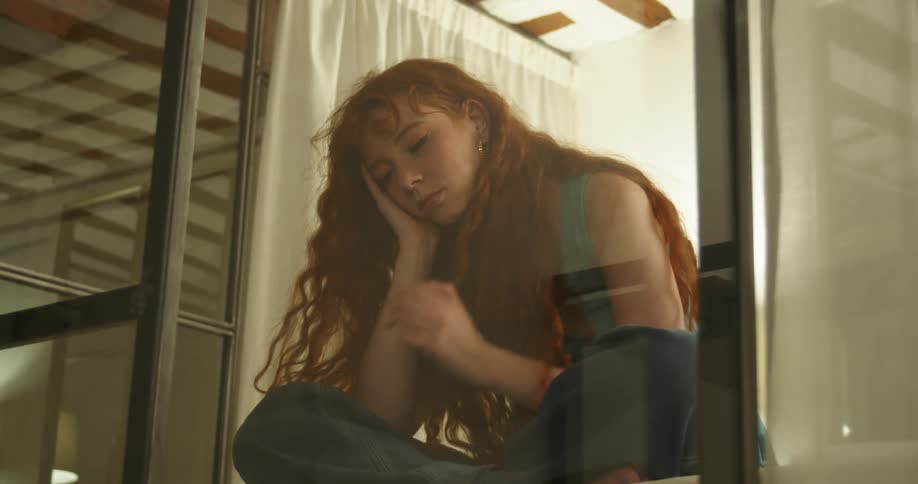 A woman is sitting indoors and looking out a window