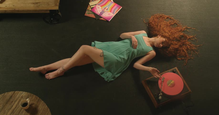 A young woman lies on the floor next to her record player and moves the needle