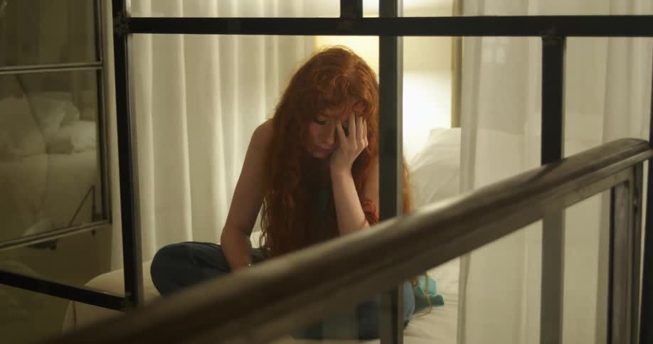 A tired looking woman rubbing her head while sitting on a bed at home