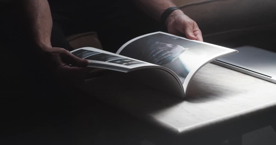 A person in a room sitting on a couch next to a table looking through a book next to a tablet