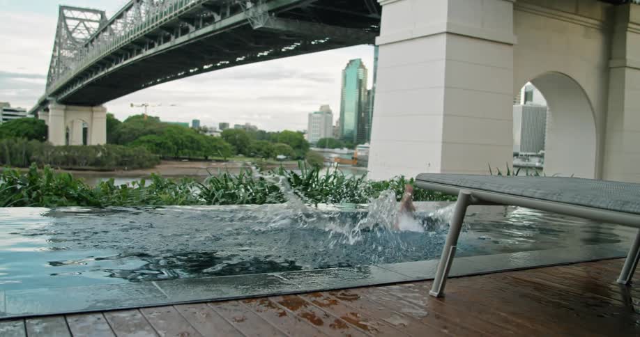 Man wearing swim trunks dives into pool near bridge support across river from city buildings