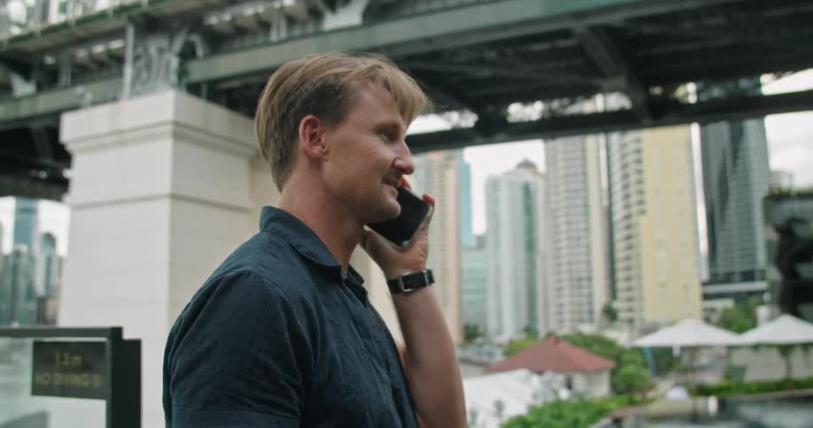 Man wearing a wristwatch talks on his smartphone while standing near a bridge in the city