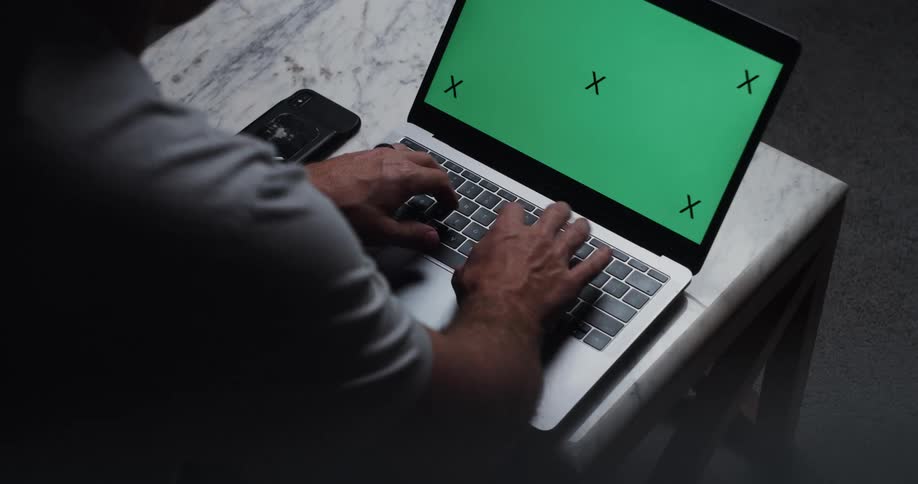 A man in a room at a desk typing on a laptop with a smart phone next to it