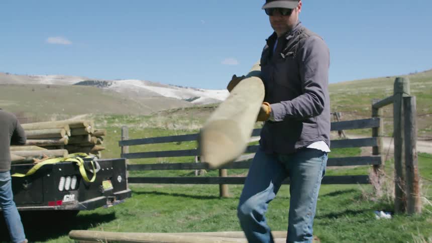 Two men unload a flatbed truck carrying fence posts on wide open ranch land in beautiful Montana mountains