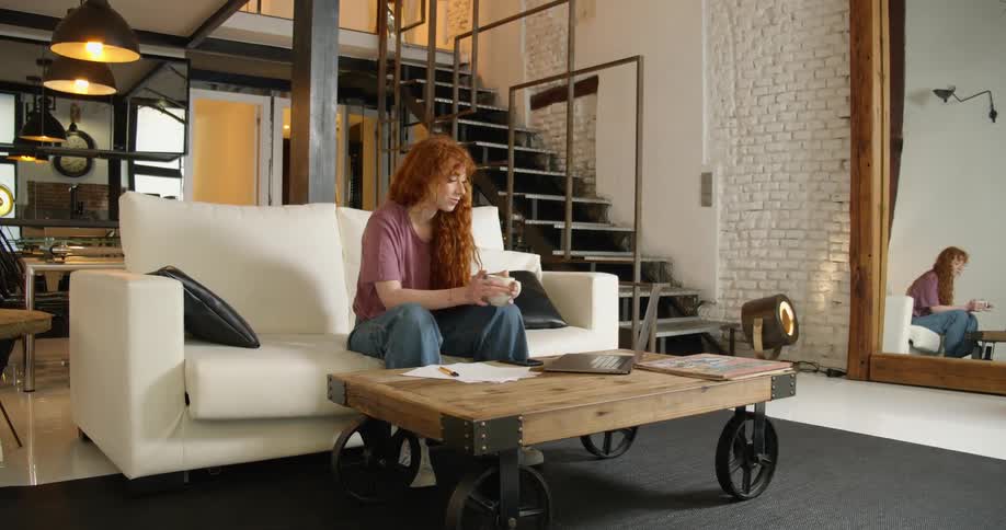 A teenage girl sits on a couch at home holding a mug and waves at her laptop as she video chats