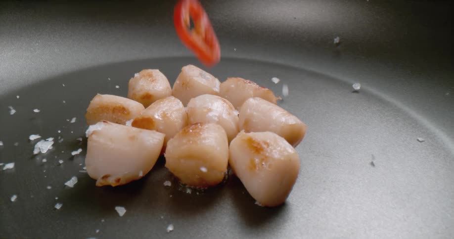 Pieces of meat sit in a pan as salt and sliced pepper fall on top