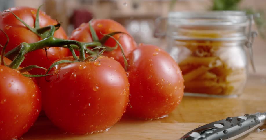 Tomatoes on a cutting board next to a knife with water droplets falling with a jar of pasta in the background