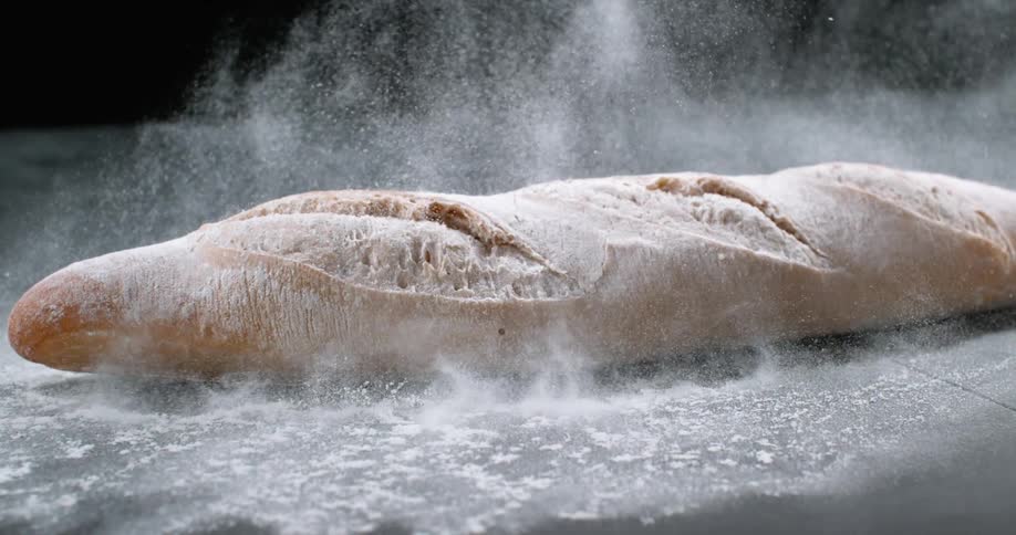 A loaf of bread covered in flour falls onto a surface in a dark room