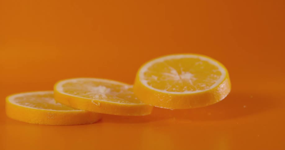 Three orange slices fall to land in spread out stack spraying juice against an orange colored background