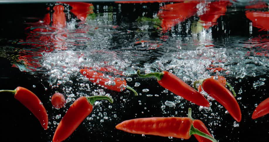 Falling bright red jalapeno peppers submerge in water against a black background