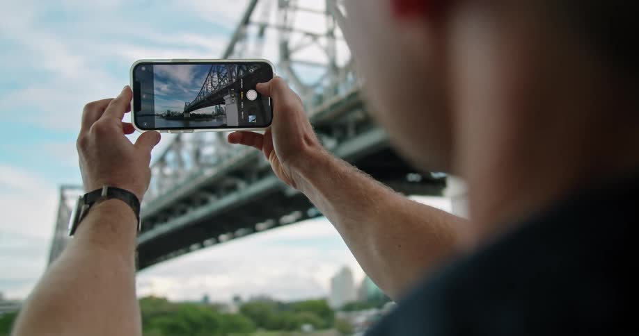A man holds up his cell phone to take a picture of a large bridge
