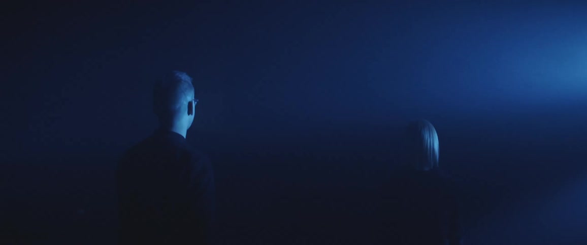 A man and woman standing outside in the dark look towards a bright light