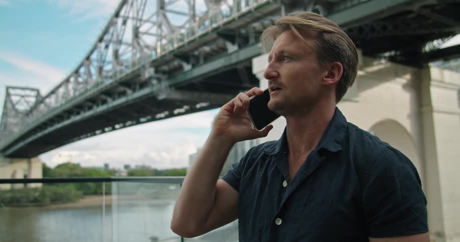 Man talks on a cell phone while standing near a bridge and a river