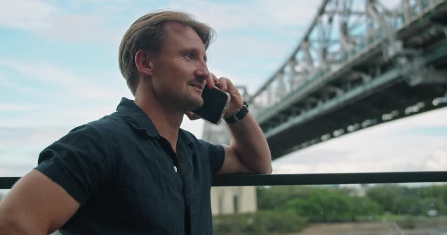 A man talking on a smart phone with a bridge the sky and clouds in the background