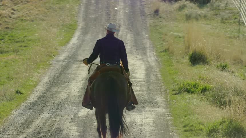 Dynamic shot of a cowboy riding down a long dusty dirt road in rural Montana
