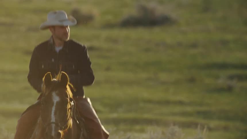 An iconic cowboy rides his horse across field while looking around in beautiful sunset light