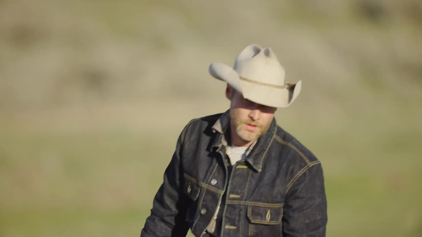 A cowboy in a denim jacket riding a horse in a grassy pasture in the far reaches the intermountain west