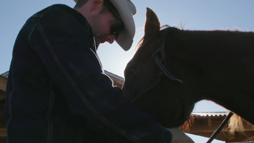 A young cowboy saddles his horse in afternoon light on a bison ranch in Montana