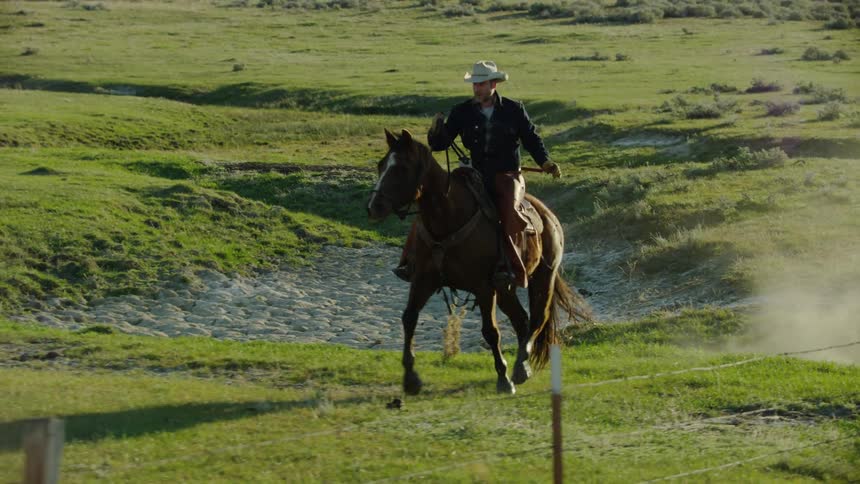 Awesome tracking slow motion shot of a brown horse kicking up dust plumes with a jean jacket young cowboy on horseback