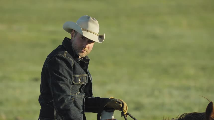 A man with a cowboy hat, denim jacket and gloves riding a horse in rural Montana at the end of long days work