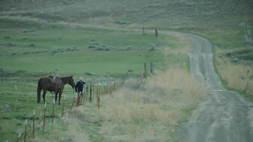 A cowboy opens a gate and leads his horse through to a rolling dirt road after the sun has set on a Buffalo ranch