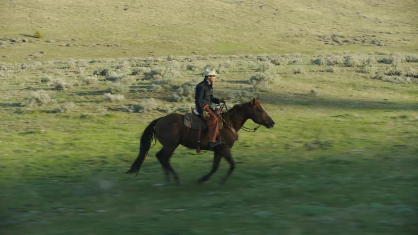 Real time high energy shot of a young cowboy riding his brown horse through an open meadow and sage brush in rural Montana