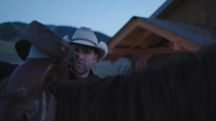 A cowboy takes the saddle off his horse after a long hard day work in rural Montana in late evening light