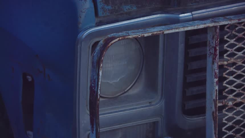 Cracked headlight on the front of a beat up ranch pickup truck that's covered in rust on a rural ranch deep in the mountains in blue hour light