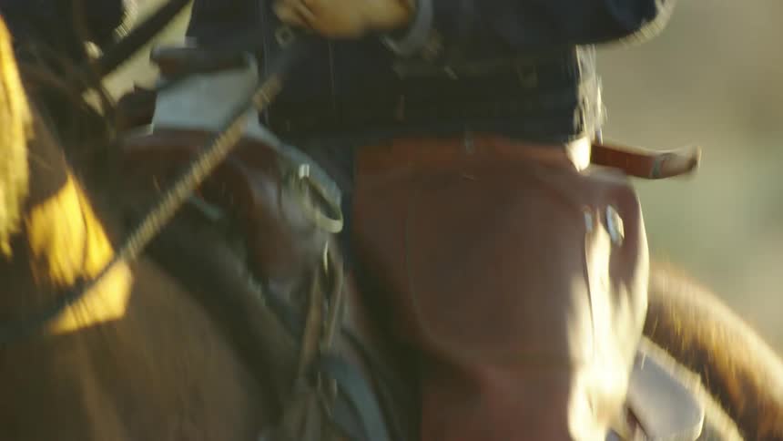 Tight shot of a cowboy wearing a cowboy hat, jean jacket and gloves riding a horse in the sunset light