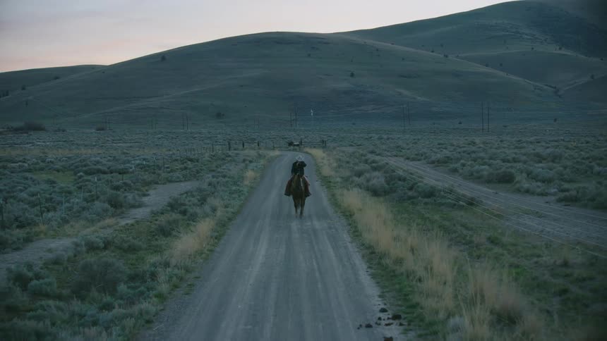 A rugged cowboy on a buffalo ranch rides his brown horse on a straight dirt road during the blue hour post sunset magic