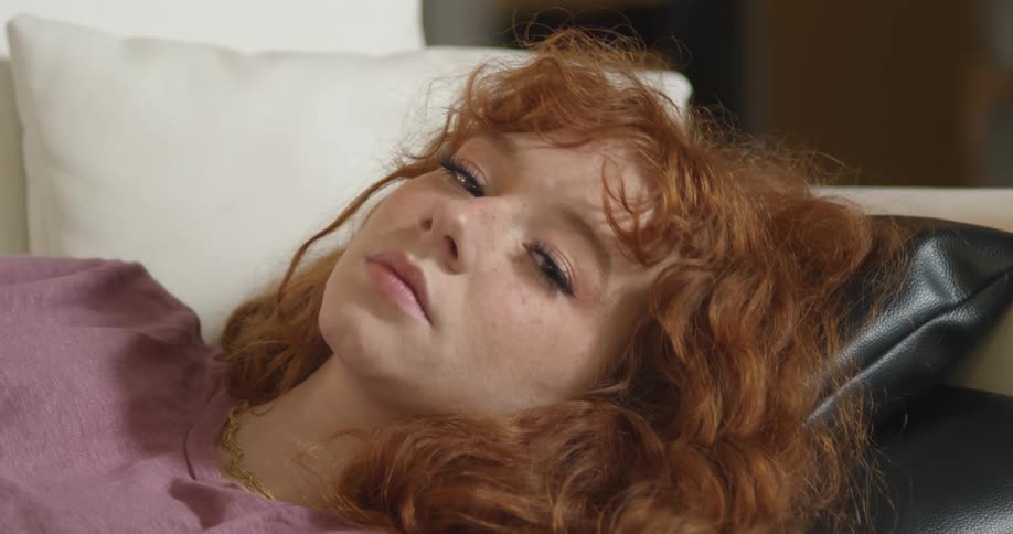 A woman in a room with a necklace laying on a couch and staring with a wall in the background