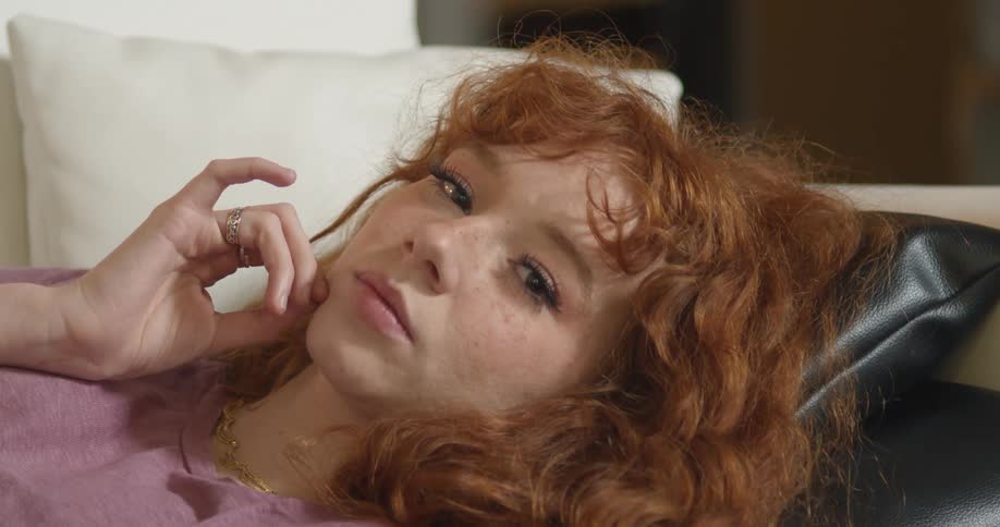 A woman with red hair and freckles reclines while looking pensive before turning to the camera