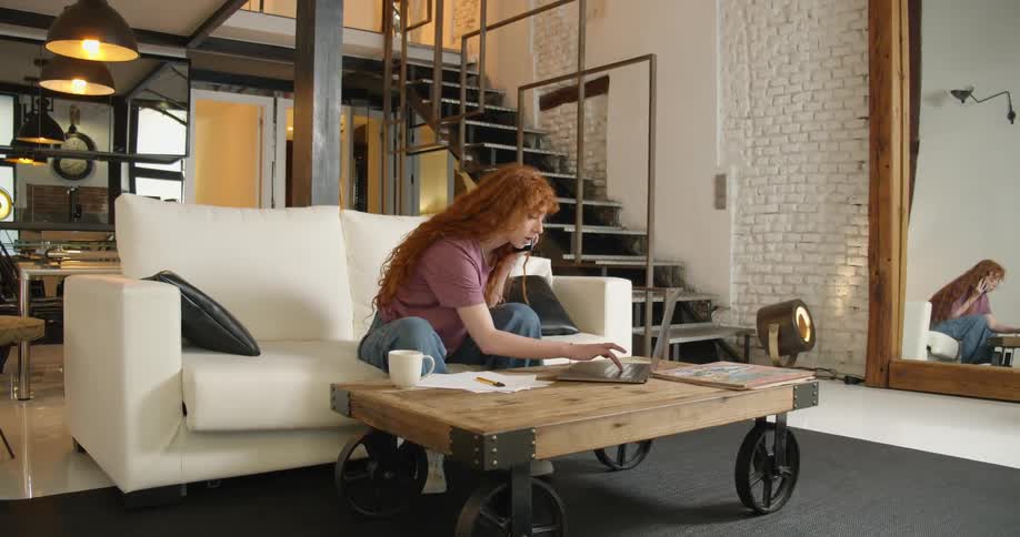 A woman sits on a couch and leans forward to type on a laptop sitting on a coffee table