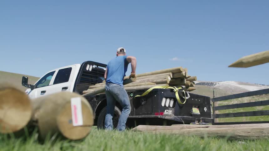 Two ranching men unload fence posts from a flatbed pickup truck in rural Montana