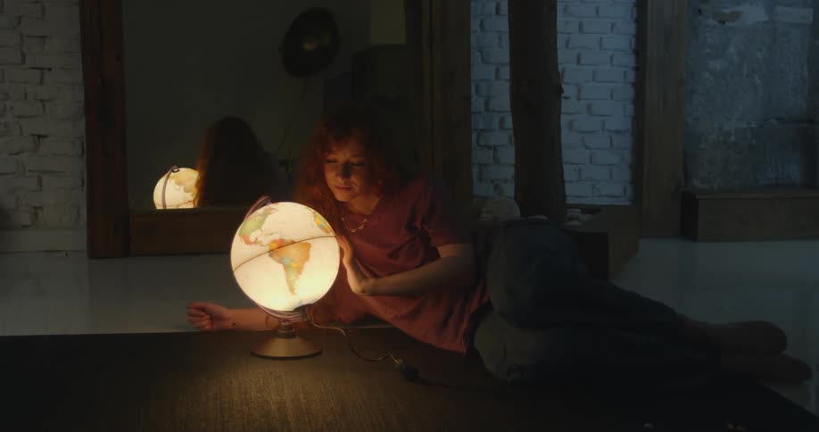 A young woman leisurely spins a a glowing globe while resting on the floor of a dimly lit room